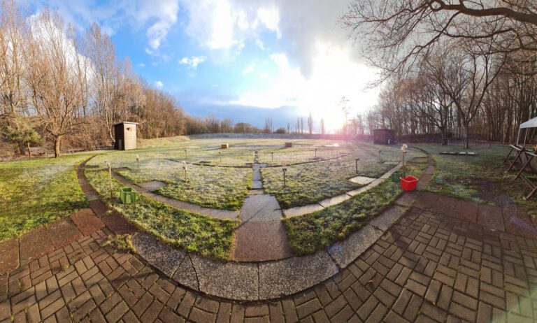 Weitwinkelaufnahme des Trapstands an einem Wintermorgen: Reif auf der Wiese, die Standplätze liegen im Halbkreis um den gepflasterten Weg im Vordergrund, die tiefstehende Sonne scheint durch kahle Bäume.