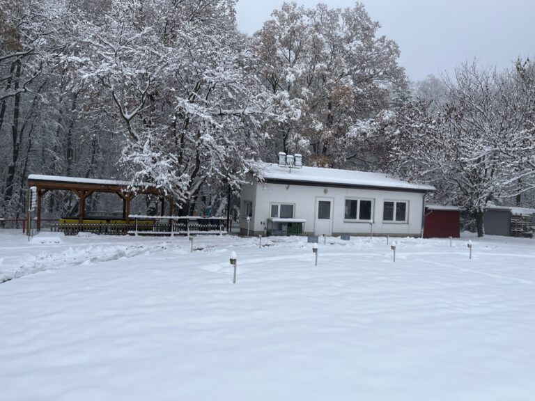 Das Schützenhaus im Schnee: ein weißer Flachbau mit drei Fenstern und einer Tür, links daneben ein offener Unterstand mit Holzdach und gelben Tischen, ringsum schneebedeckte Bäume.