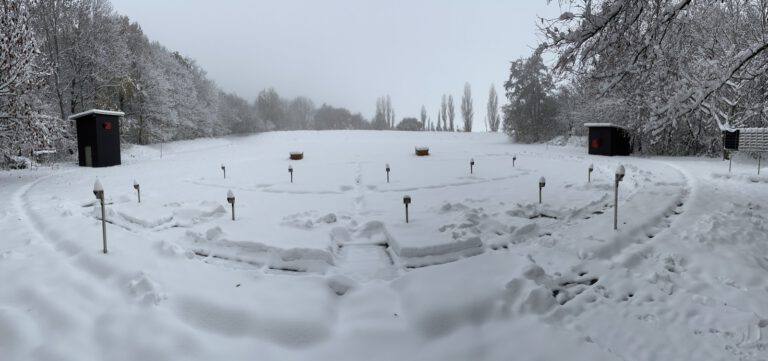 Panorama des Stands nach Neuschnee: aus der weißen Fläche ragen nur die Markierungssäulen der Standplätze und zwei Wurfmaschinenbunker, links und rechts je ein dunkles Holzhäuschen, im Hintergrund Nebel.