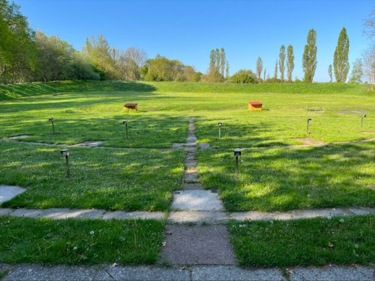 Die Trapanlage im Frühling: fünf Standplätze aus Betonplatten im Gras, dahinter auf der Wiese zwei Wurfmaschinenbunker mit rot und gelb gedeckten Dächern, am Horizont Pappeln unter blauem Himmel.